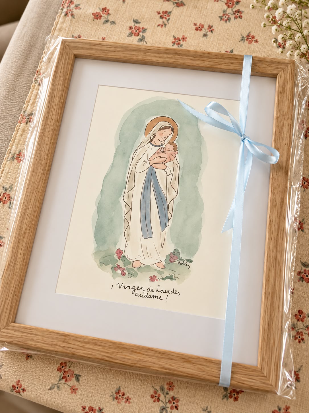 Fotografía de una acuarela enmarcada de la Virgen con el Niño, envuelta con un lazo azul y las palabras Virgen de Lourdes, cuídame
